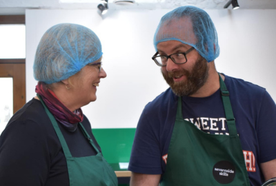 A person we support and staff member in the kitchen wearing aprons and hair nets preparing food and talking in Taurus Crafts, Severnside Skills vocational courses for adults with a learning disability